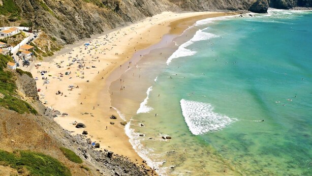 Uma praia costeira com um penhasco rochoso, areal arenoso e águas turquesa cristalinas.