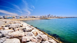 Un paysage urbain côtier avec une rive rocheuse, une plage de sable et une rangée de bâtiments le long du bord de l’eau.