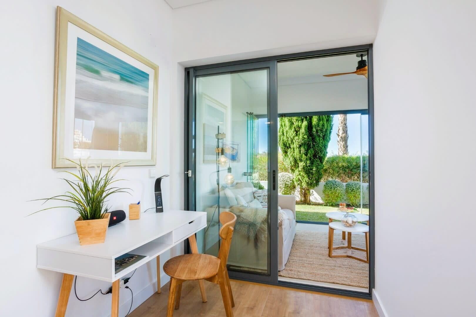 Een moderne woonkamer met een wit bureau, een potplant en een schuifdeur die leidt naar een balkon met uitzicht op groen.