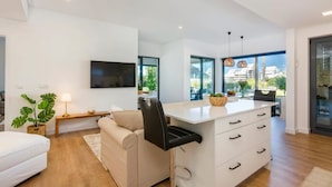 Un salon moderne comprenant un canapé, un téléviseur fixé au mur, un coin repas avec une table et des chaises, et une cuisine dotée d’un plan de travail et de meubles blancs.