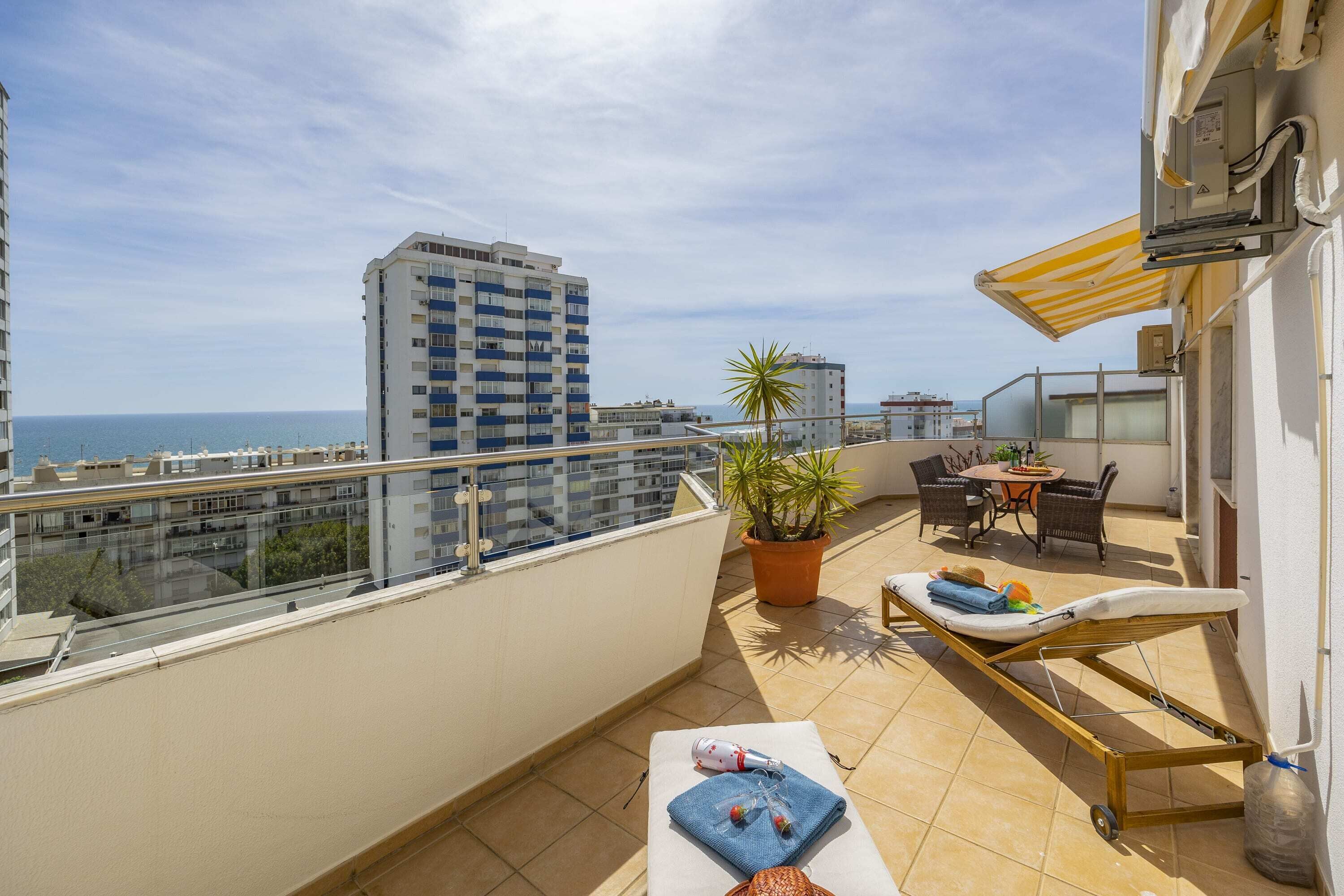 Een balkon met een ligbed, een tafel met stoelen, een plant in pot en uitzicht op zee en gebouwen.