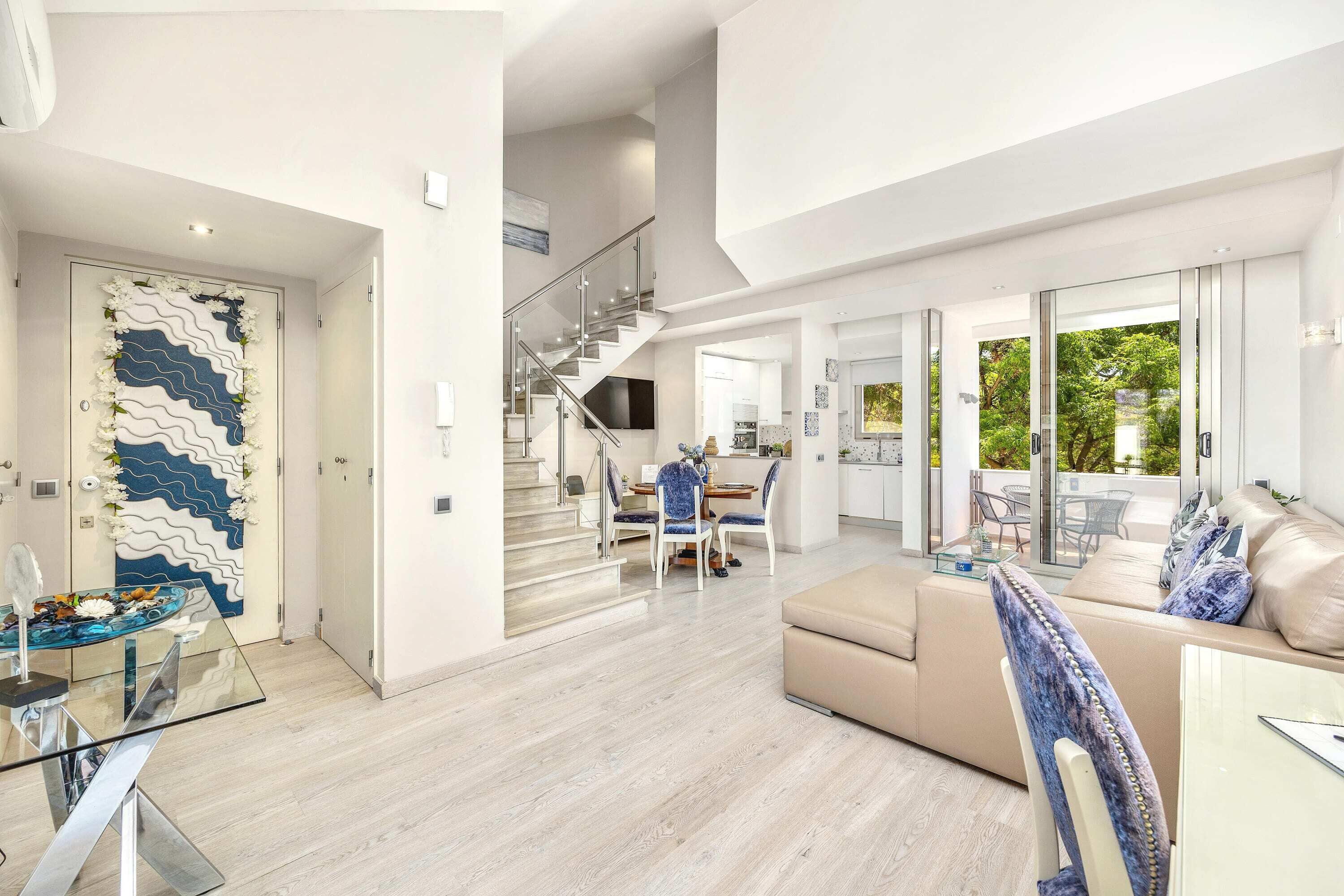 A spacious living area with a modern staircase, a dining area with a glass table, and a large window offering a view of greenery.