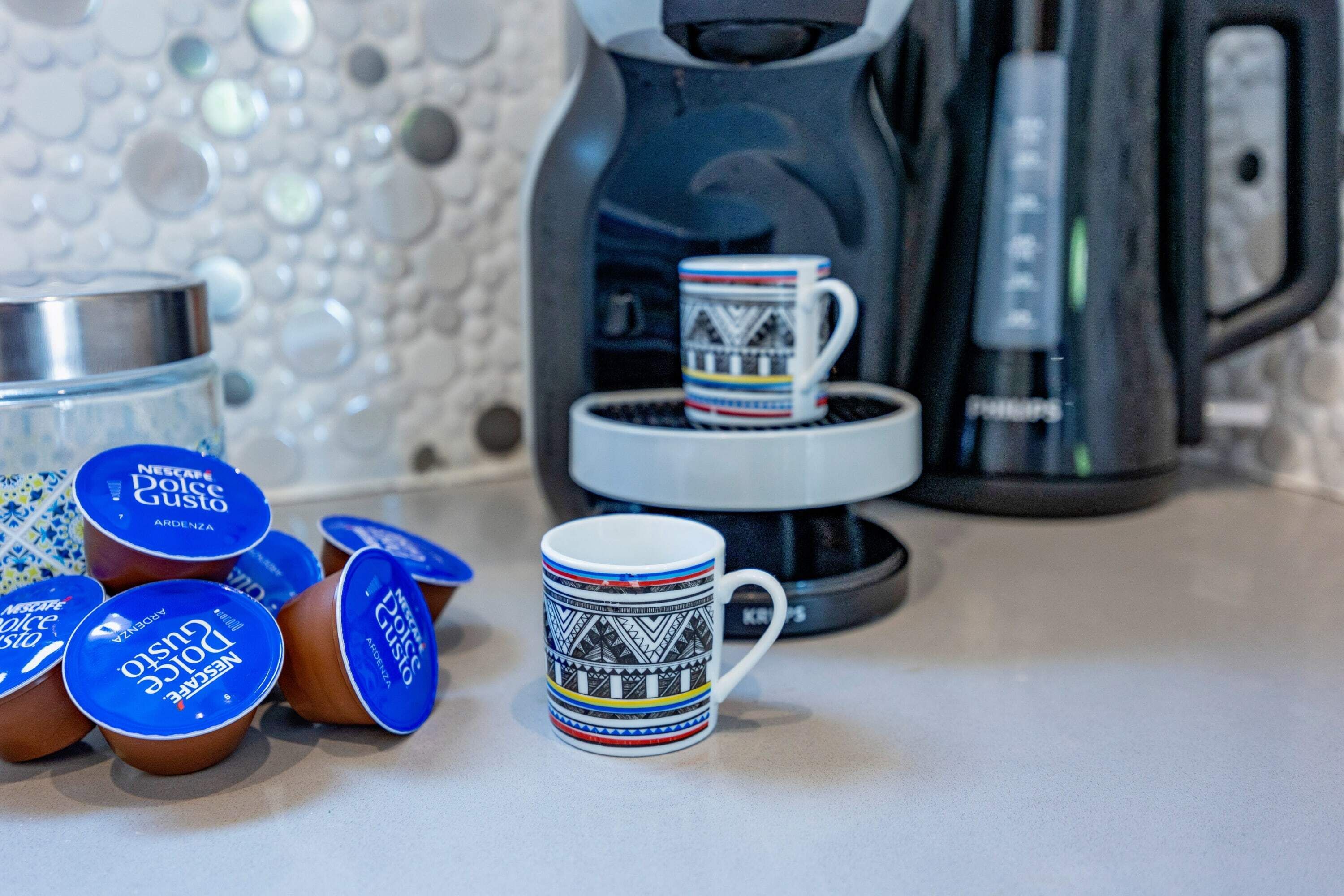 A kitchen counter with a coffee maker, a mug, and several containers of Dolce Gusto coffee.