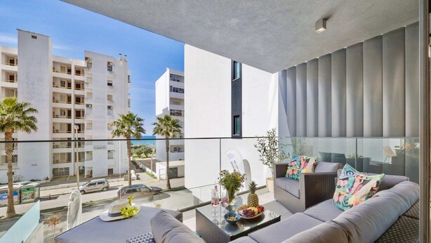 Ein Balkon mit einer Couch, einem Glastisch mit Früchten und Getränken sowie Blick auf ein modernes Gebäude und Palmen.