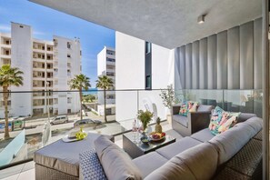 Un balcon avec un canapé, une table en verre sur laquelle sont disposés des fruits et des boissons, et une vue sur un immeuble moderne et des palmiers.