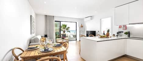 A modern kitchen with a central island, a dining area with a wooden table and chairs, and a view of the ocean through large windows.