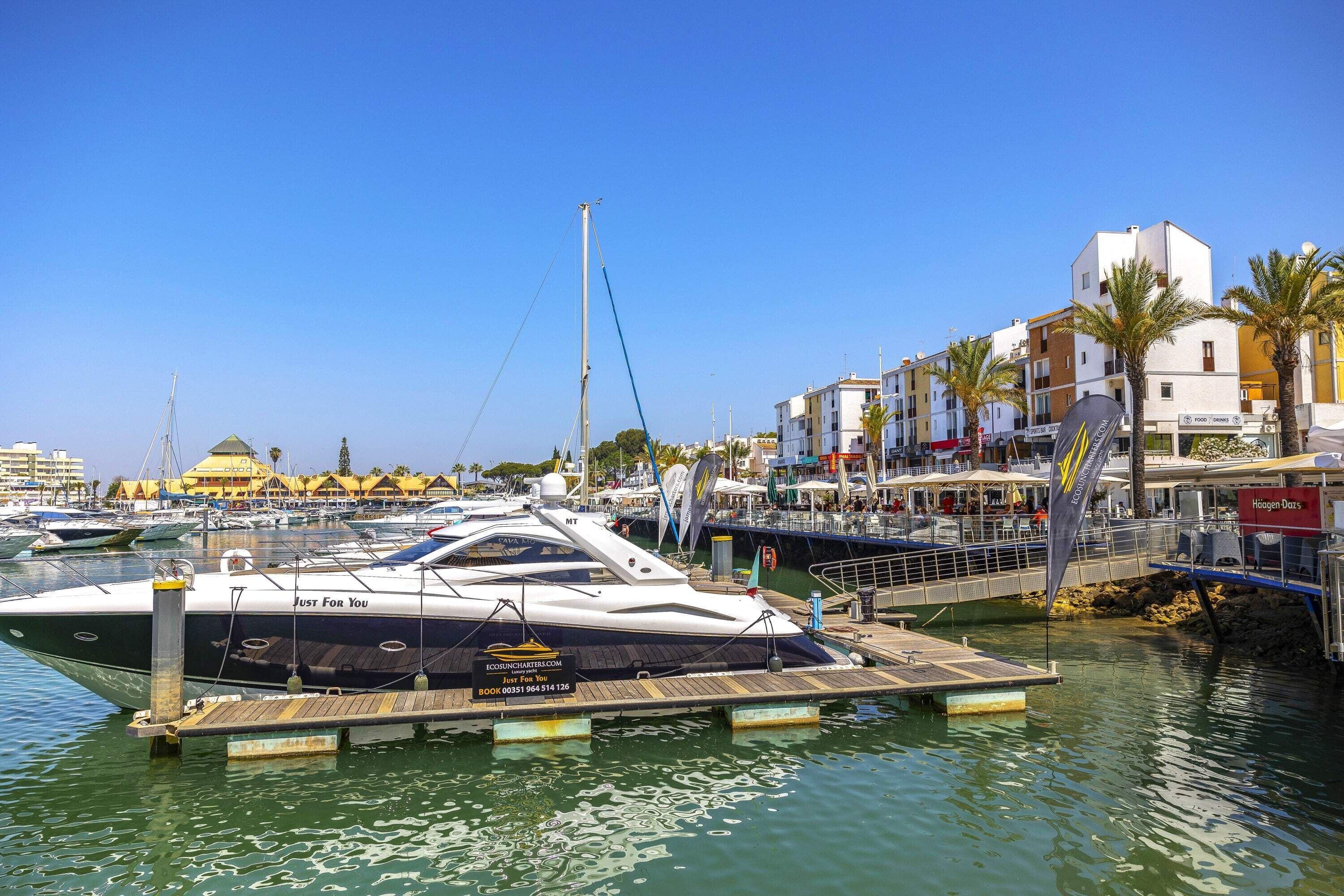 A marina with boats docked, a wooden pier, and buildings with shops and restaurants in the background.
