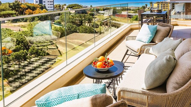 Un balcon agrémenté de meubles en osier, un panier de fruits et une vue sur la mer et les bâtiments.