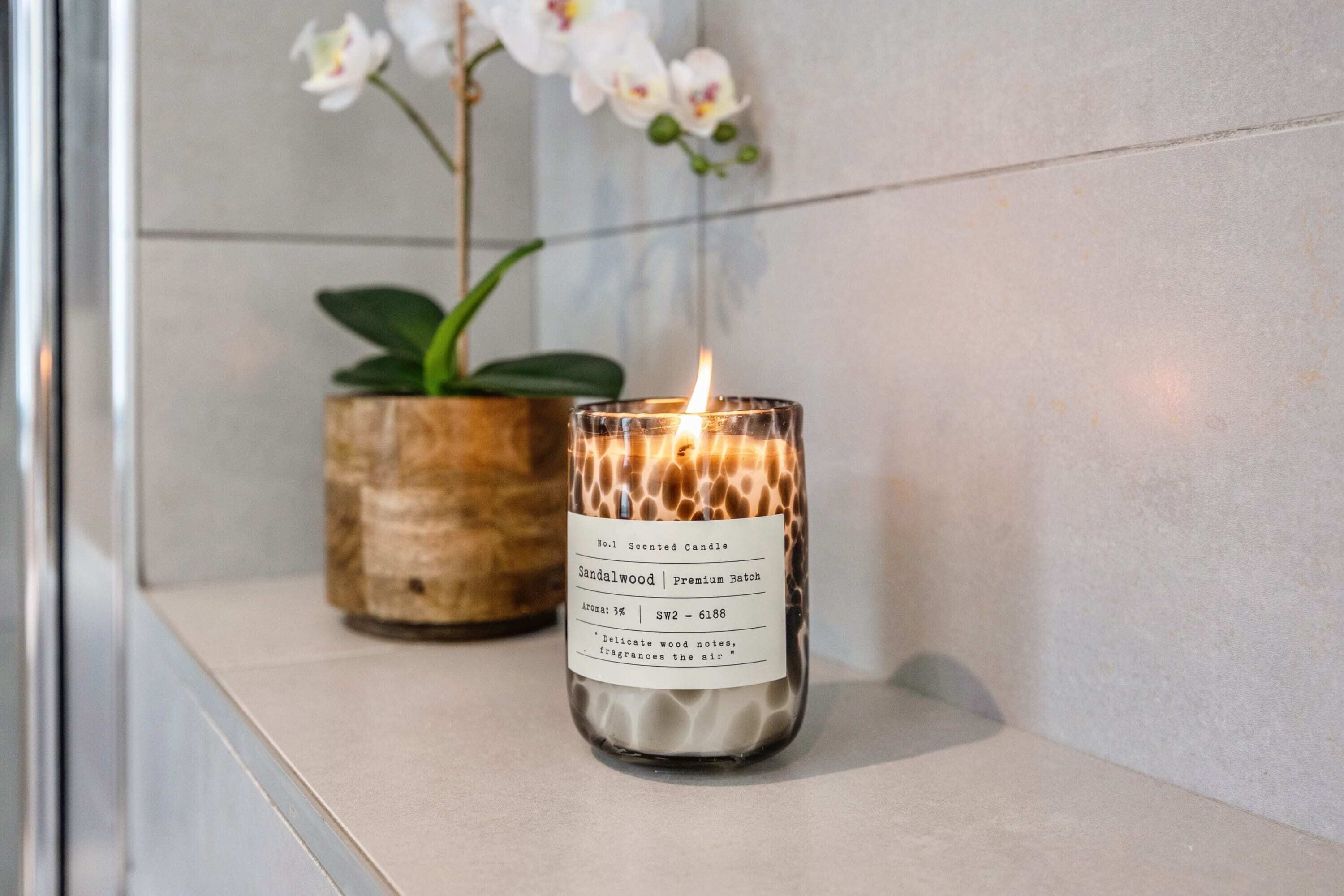 A scented candle labeled 'Sandalwood Premium Batch' with a visible flame, placed on a shelf next to a potted plant.