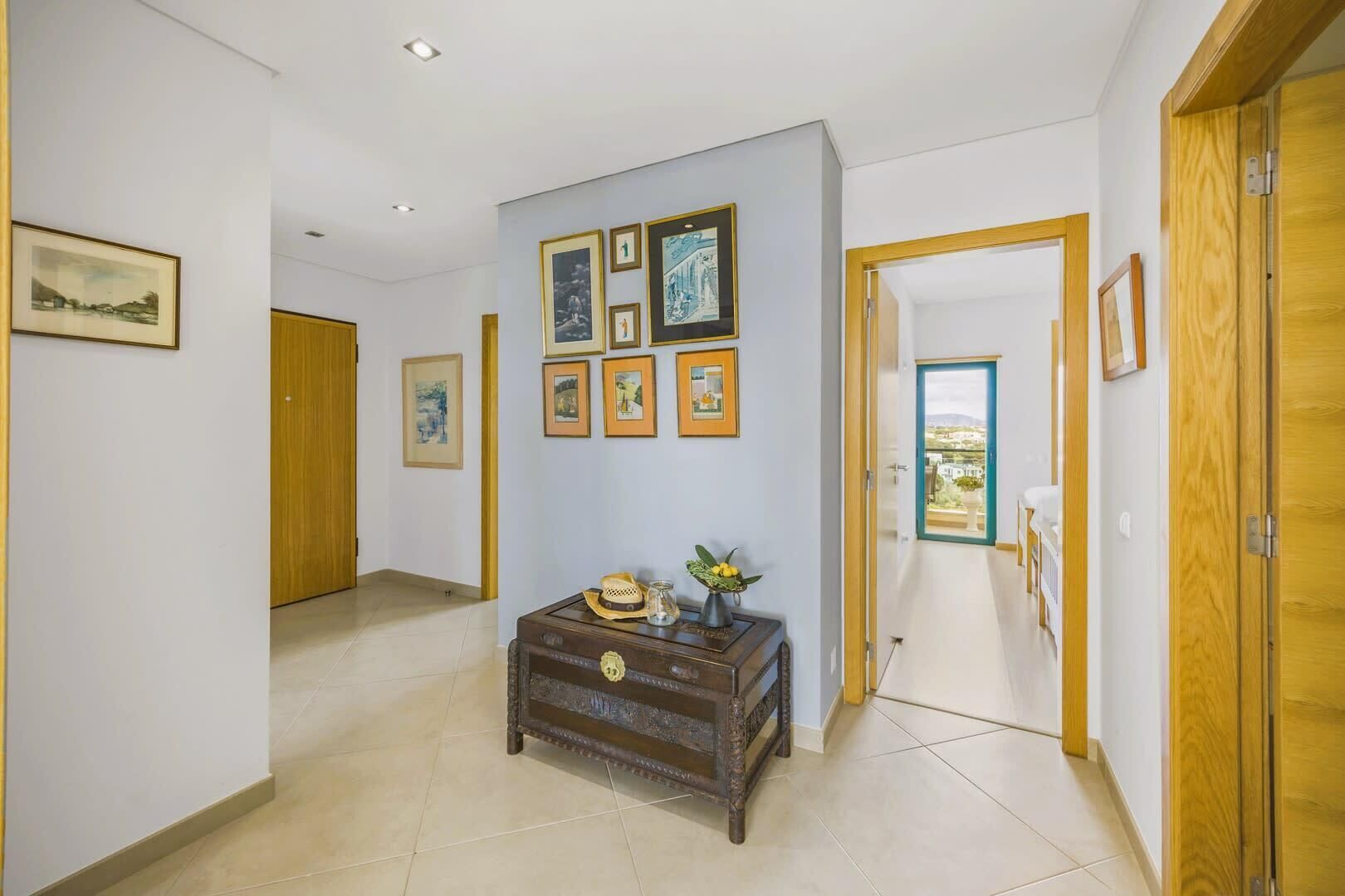 A hallway with framed pictures on the wall, a wooden chest, and a view through an open door.