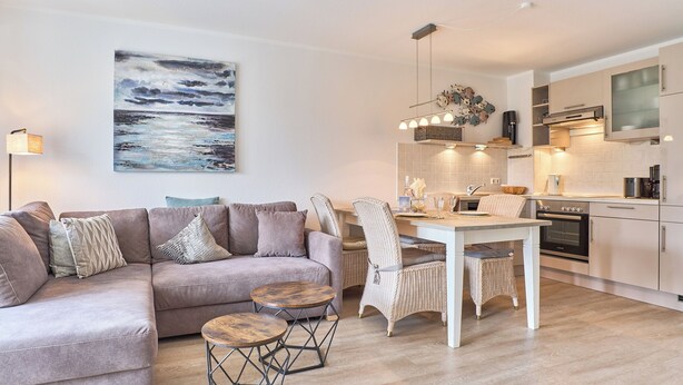 A modern living room with a sofa, two round tables, and a dining table with chairs. A kitchen with white cabinets and a stove is visible in the background. - Hansa-residenz II Appartement 9 (Scharbeutz)