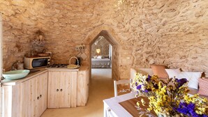 Un interior rústico de piedra con una zona de cocina, una mesa de comedor con flores, y un dormitorio acogedor con cama y almohadas.