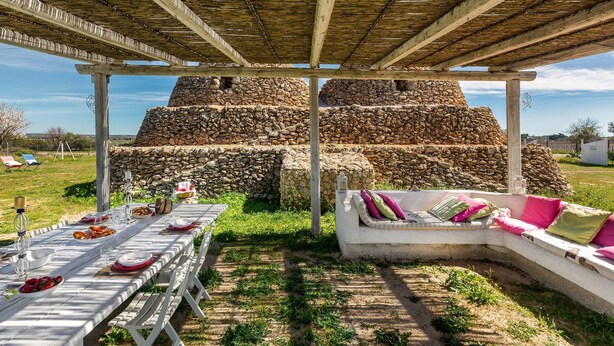 Un área de comedor al aire libre con un muro de piedra de fondo, un espacio de estar cubierto y una vista despejada del paisaje circundante.