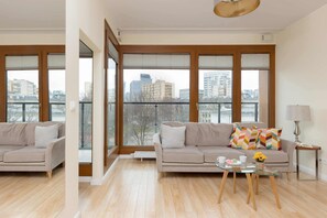 Een moderne woonkamer met een beige bank, een glazen salontafel en uitzicht op de stad door grote ramen.