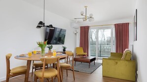 A modern dining area with a round wooden table, yellow chairs, and a flat-screen TV mounted on the wall.