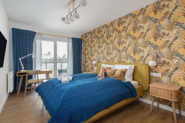 A cozy bedroom with a double bed, blue bedding, and a decorative yellow-patterned wallpaper. The room is bright and modern.