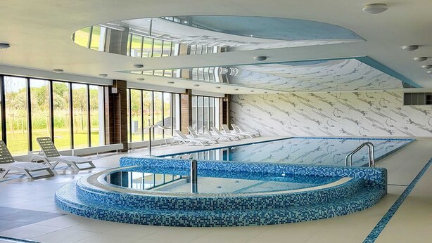 A modern indoor swimming pool with a blue mosaic design, surrounded by lounge chairs and large windows offering a view of the outdoors.