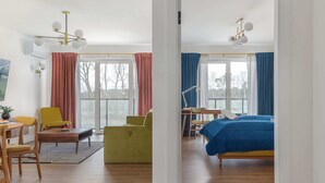 A modern hotel room with a wooden floor, a bed with a blue duvet, a desk with a chair, and a dining area with yellow chairs.