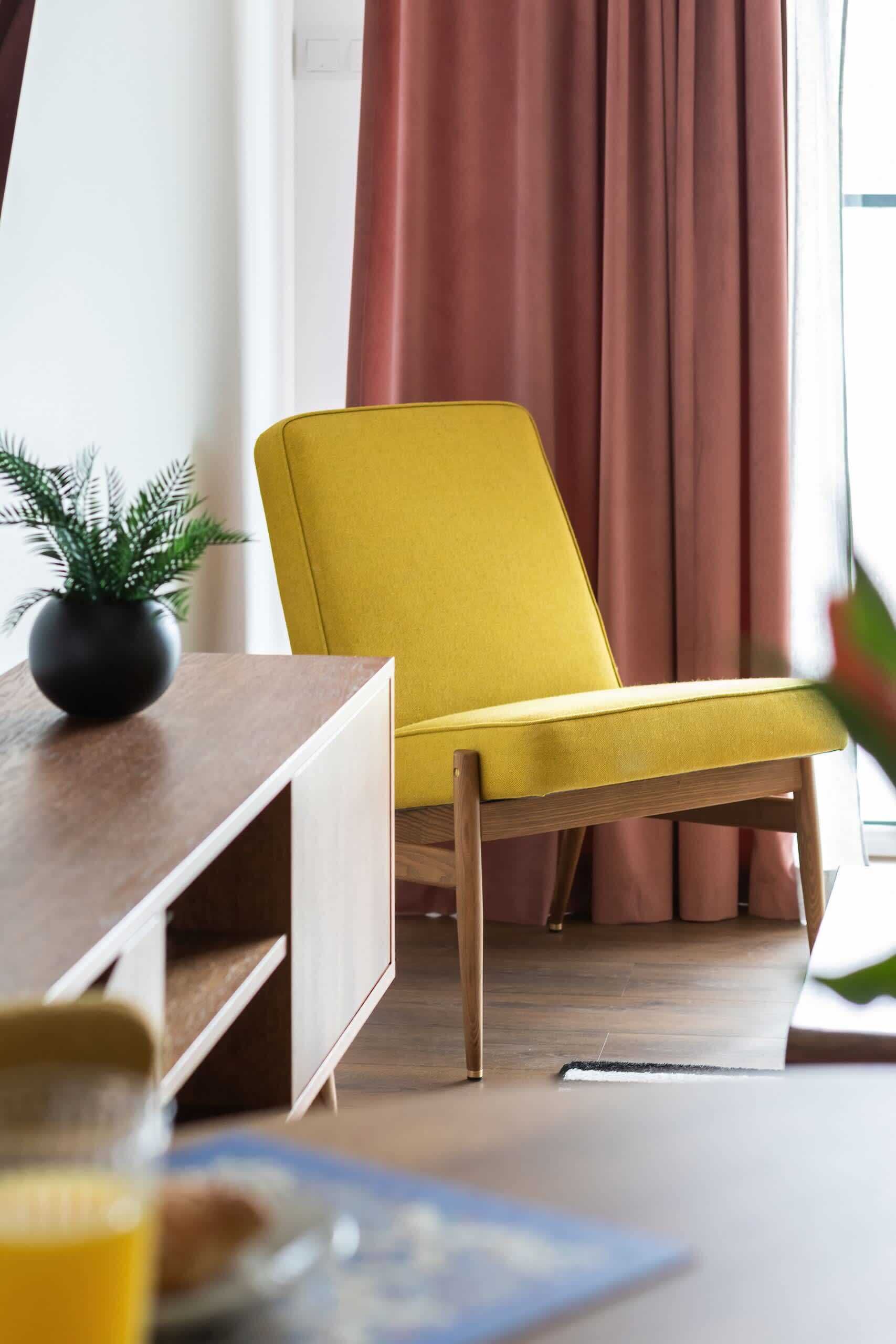 Un fauteuil jaune dans une pièce dont le sol est en bois, une petite plante posée sur une table et une fenêtre avec des rideaux.