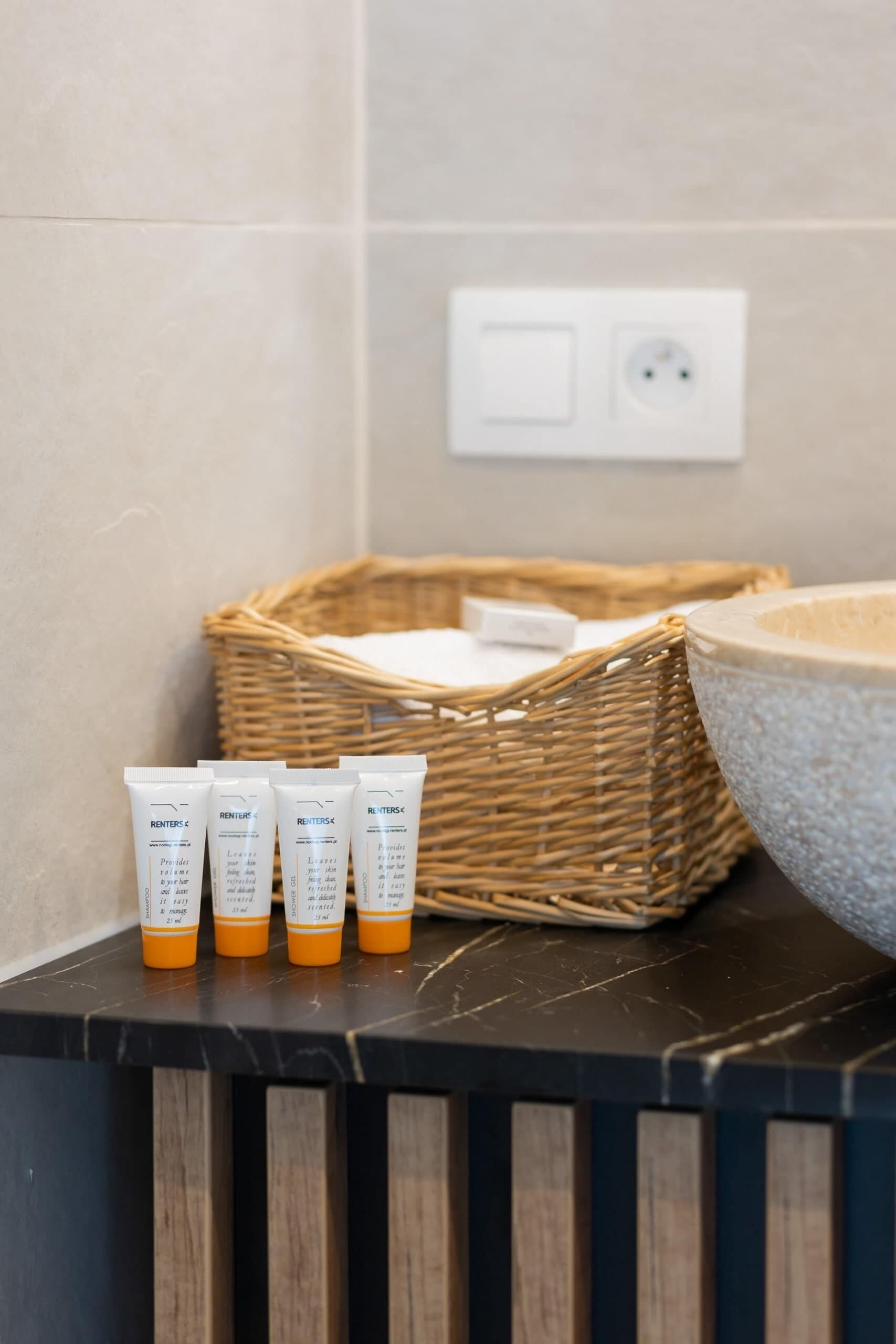Une salle de bain avec un grand lavabo en pierre, un panier en osier et trois flacons de produits de toilette posés sur un plan de toilette foncé.