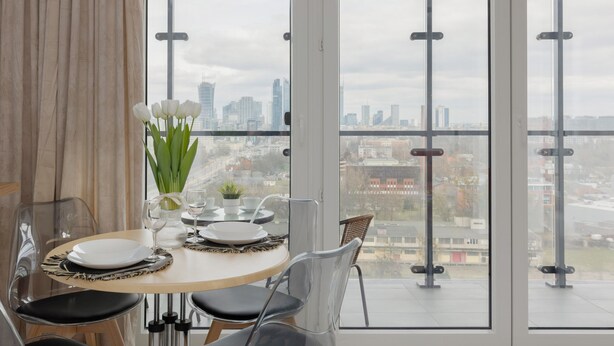 A dining area with a table set for four, large windows with a city view, and a curtain on the left. - Modern Studio with Balcony | Parking | Close to EXPO XXI (Warszawa)