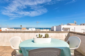 Una terraza en la azotea con una mesa y sillas, una maceta blanca con cactus y una vista despejada al mar y edificios al fondo.