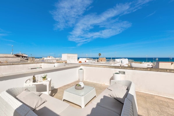 Una terraza en la azotea con mobiliario de mimbre blanco, una jardinera central con flores y vistas despejadas al mar y al paisaje urbano.