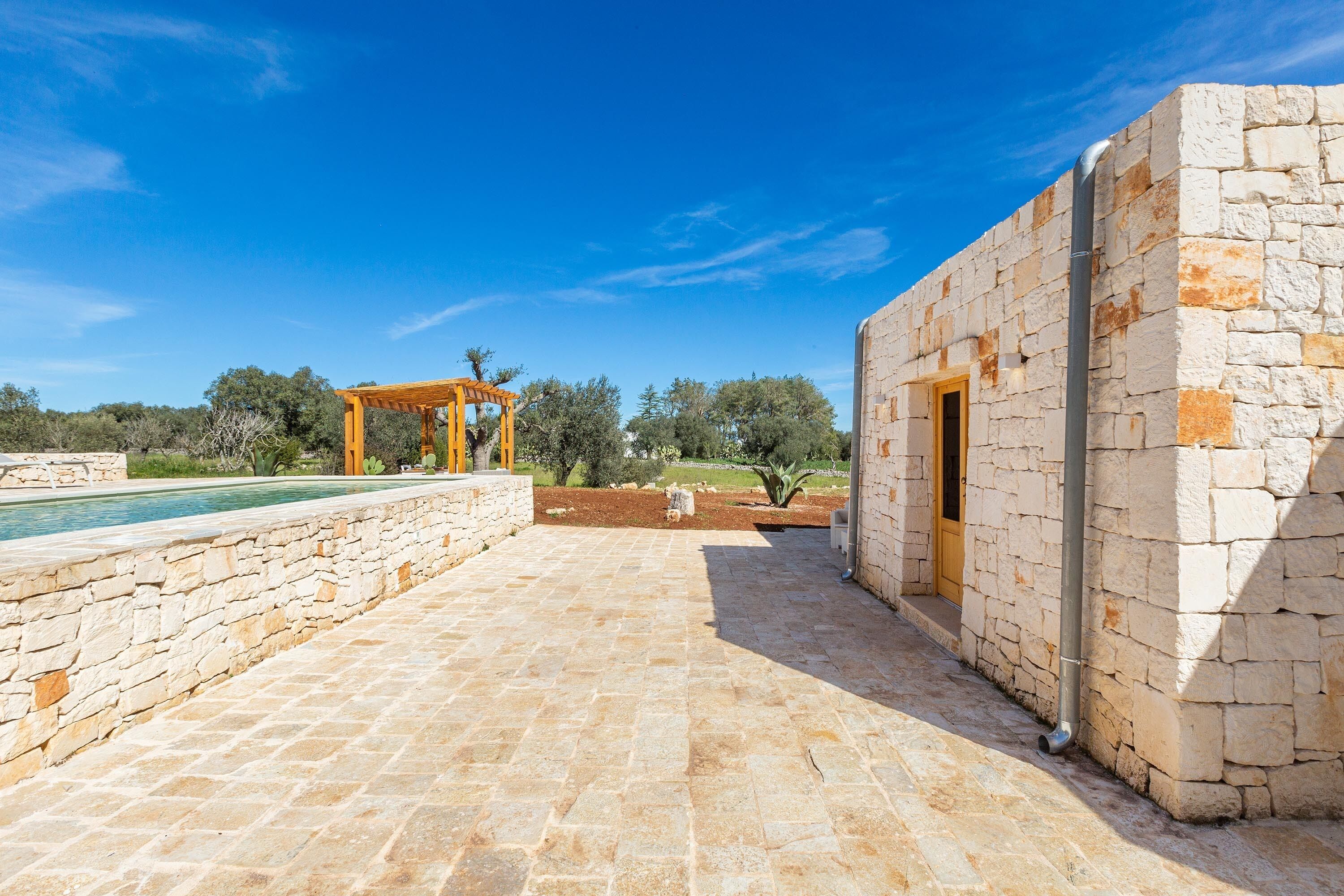 Um pátio de pedra com pérgola, piscina e um edifício de pedra com porta.
