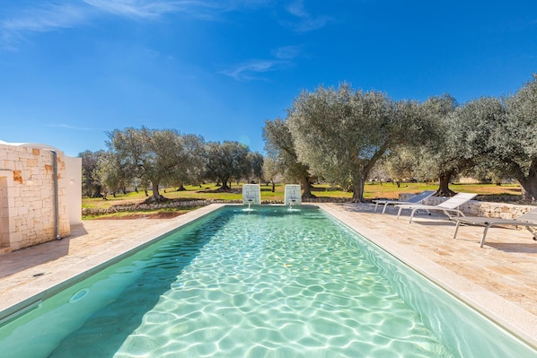 A rectangular swimming pool with clear water, surrounded by a stone deck and lounge chairs.