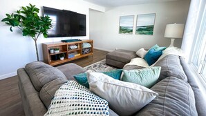 Una sala de estar con un sofá gris, un televisor de pantalla plana, un mueble de televisión de madera y una planta.
