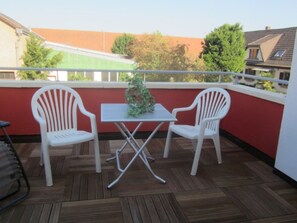 Ein Balkon mit zwei weißen Plastikstühlen und einem kleinen Tisch, Holzboden und einer roten Wand.