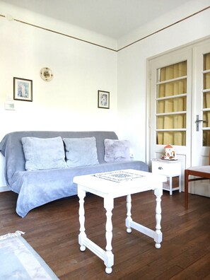 Ein Zimmer mit grauem Sofa, einem weißen Tisch mit gemustertem Tischläufer, einem Holzstuhl, einem weißen Nachttisch und einer Tür mit Fenster.