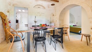 A dining area with a rustic stone wall, a wooden ceiling beam, and a table set with chairs. - 3224 Trulli Almaviva by Perle di Puglia (Cisternino)