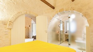 A room with a yellow bed, stone walls, and an arched doorway leading to a bathroom. - 3224 Trulli Almaviva by Perle di Puglia (Cisternino)