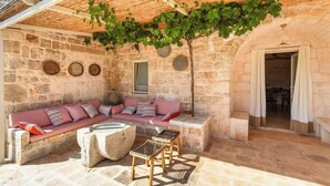 A stone-walled outdoor seating area with a cushioned bench, a small table, and a pergola with vines. - 3224 Trulli Almaviva by Perle di Puglia (Cisternino)
