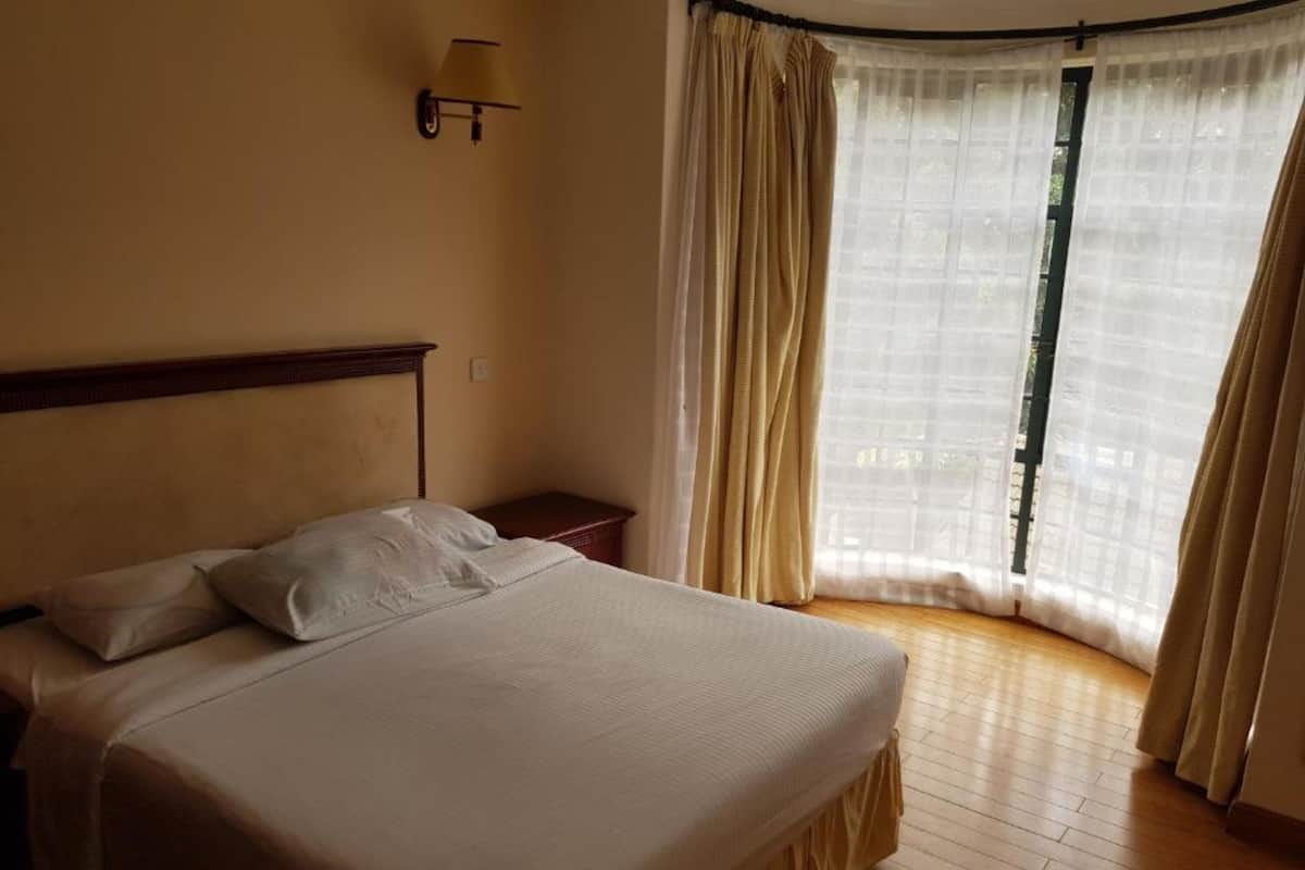 Ein Schlafzimmer mit Bett, Nachttisch, Wandlampe und Fenster mit durchsichtigen Vorhängen.