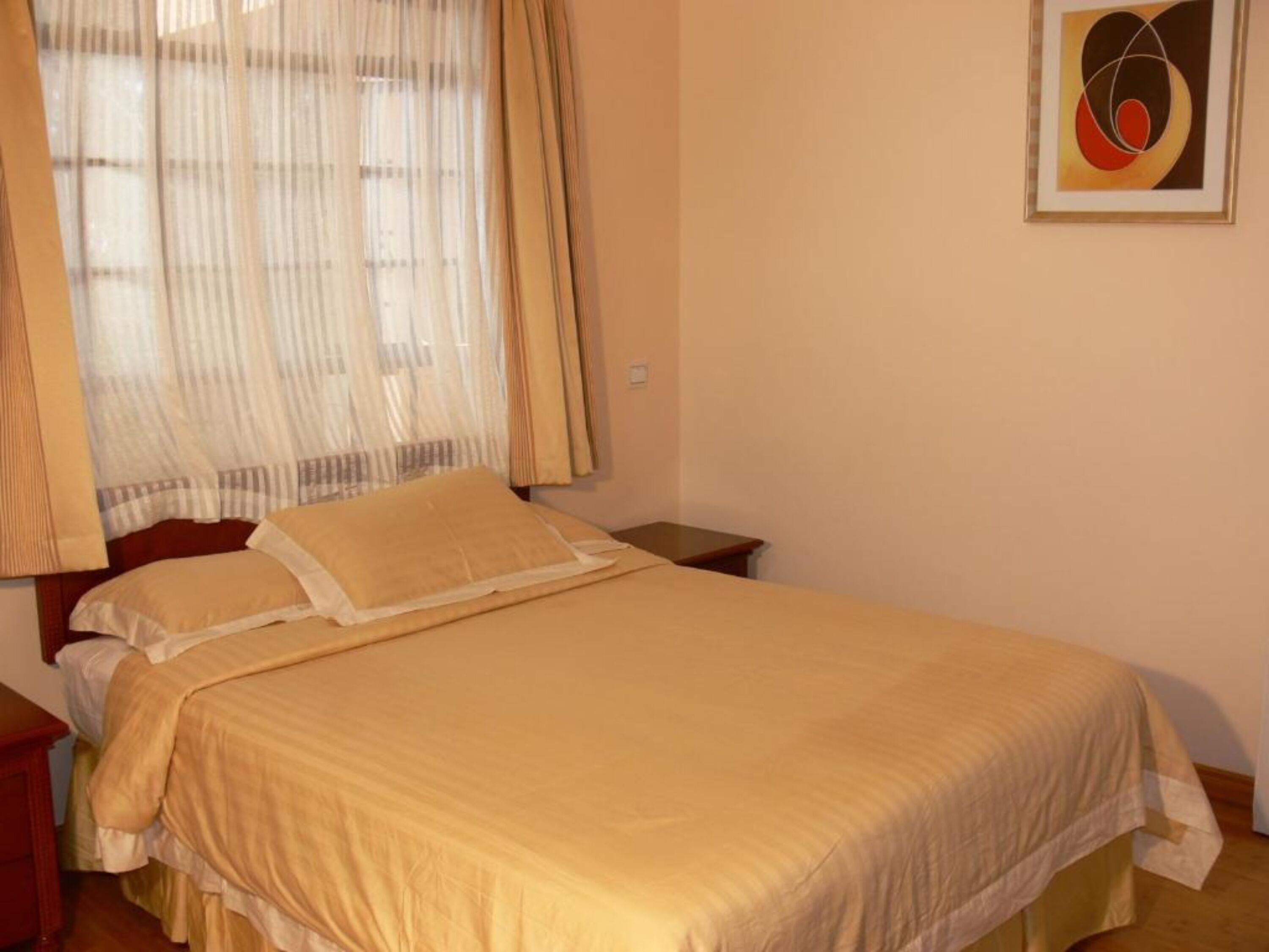 Una cama bien hecha con una colcha color beige y almohadas, una mesita de noche y una ventana con cortinas translúcidas.