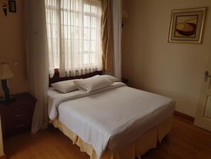 Un dormitorio con cama, mesita de noche, lámpara, ventana con cortinas e imagen colgada en la pared.