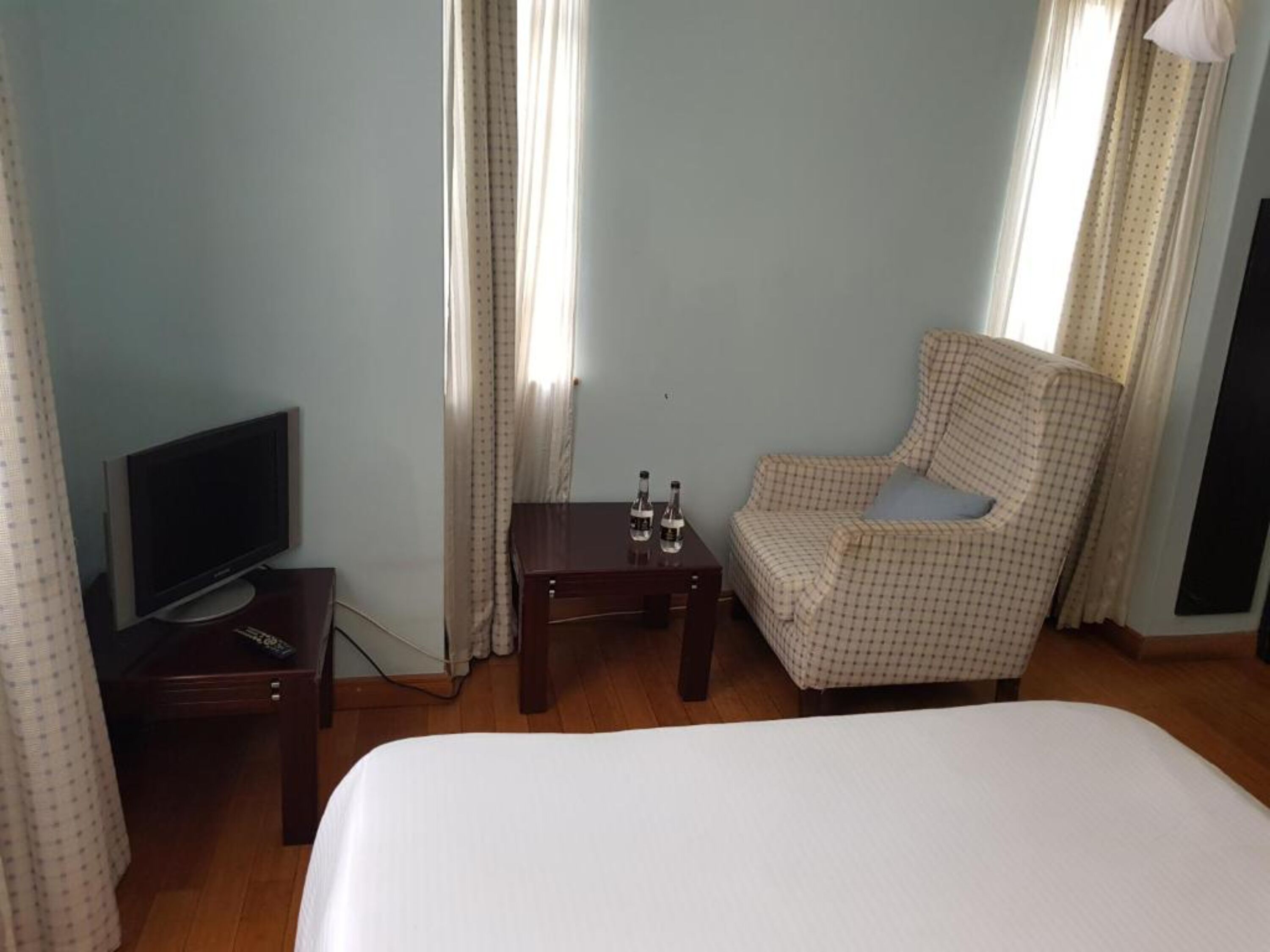 Una habitación de hotel con una cama, televisión, una mesita con botellas, una silla y cortinas.