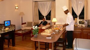 A hotel room with a dining area, a sofa, a TV, and a person in chef attire preparing food. - Natural Oak Apartments (Nairobi)