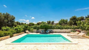 Une piscine rectangulaire bordée de pierres, entourée de chaises longues et d’une table, le tout se fondant dans un décor de verdure et sous un ciel dégagé.