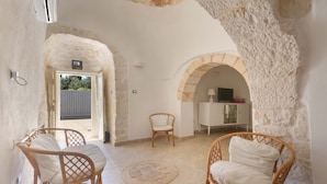 Une pièce avec deux fauteuils en osier, une petite table et un meuble blanc. Une porte donne sur l’extérieur, et les murs ont un revêtement texturé.