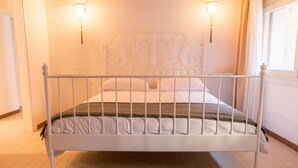 Un letto in metallo bianco con testiera decorativa, due lampade a parete e una finestra con tende.
