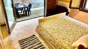 Un dormitori amb un llit, una taula petita amb cadires, un radiador i vista a un espai exterior amb una paret de maó i una taula.