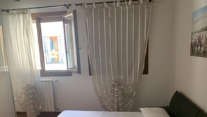 Un dormitorio con una cama, una ventana con cortinas, un radiador y un cuadro en la pared.