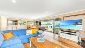 A spacious living room with a blue sofa, wooden coffee table, and a television displaying a beach scene. - Tanapaser – spacious centrally located home with fireplace (Currarong)