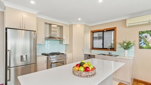 A modern kitchen with stainless steel appliances, a central island with fruit, and a window with blinds. - Tanapaser – spacious centrally located home with fireplace (Currarong)