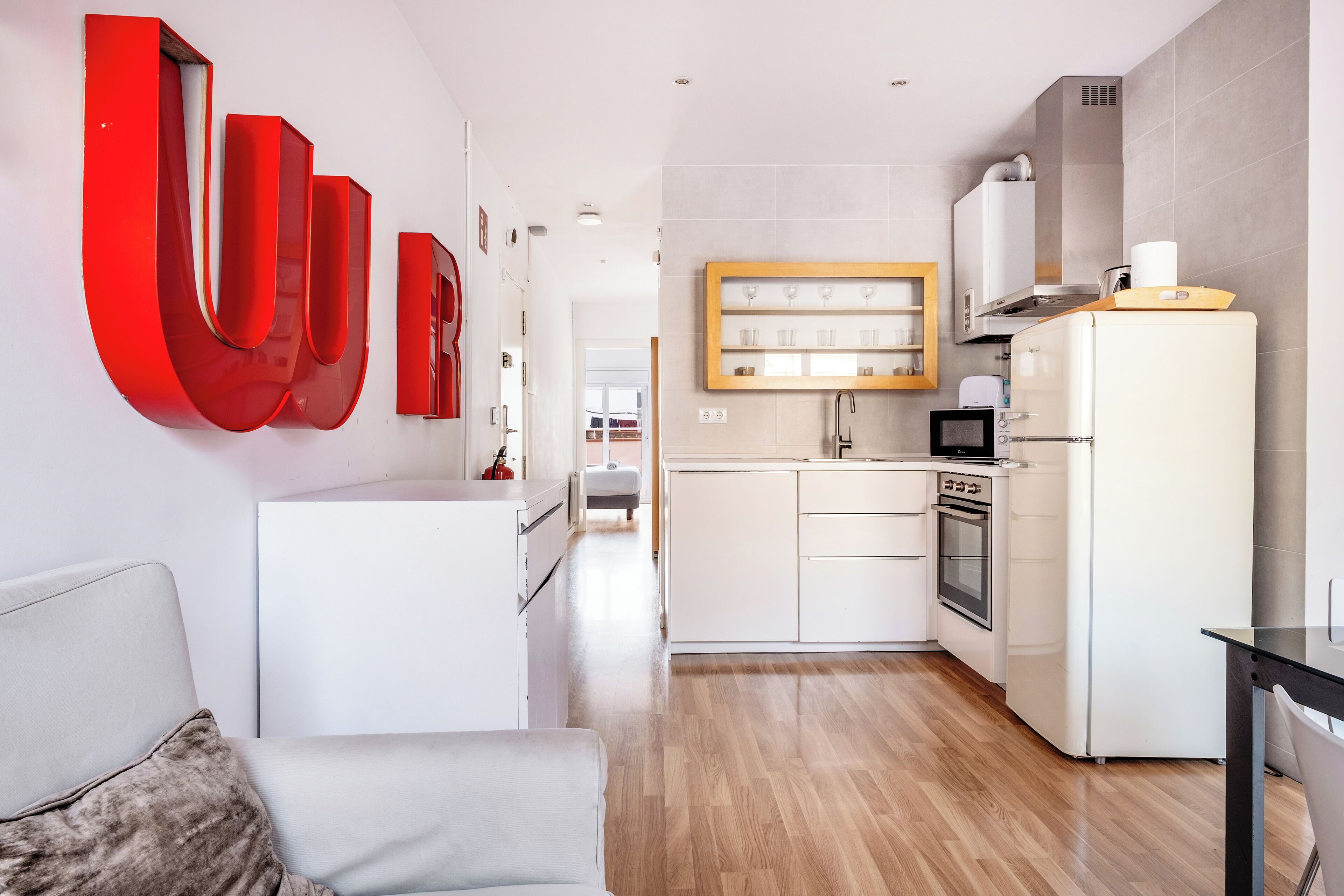 Une cuisine moderne avec des armoires blanches, un four encastré et un four à micro-ondes. Une étagère en bois est fixée au-dessus de l’évier.