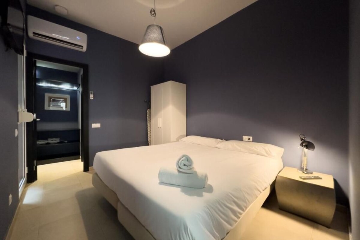 Una cama bien hecha con una toalla enrollada, una lámpara de noche y un armario en una habitación con paredes oscuras y un equipo de aire acondicionado.