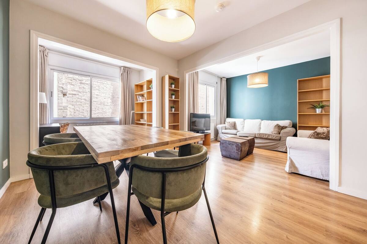 A modern living room with a wooden dining table, green upholstered chairs, a white sofa, and built-in wooden shelving.